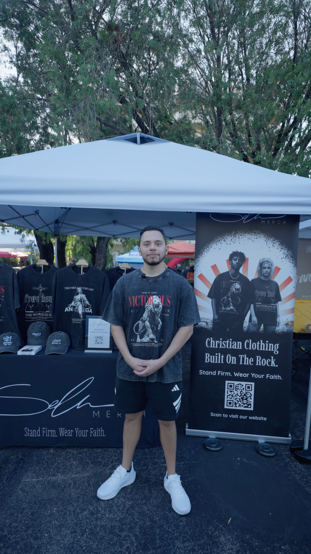 Sela Founder, Jesus Marquez, stands in front of a white pop-up canopy at an outdoor market, wearing a black graphic t-shirt, black shorts, and white sneakers. A black banner on the right reads 'Christian Clothing Built On The Rock' with a QR code, and merchandise is displayed on the left.